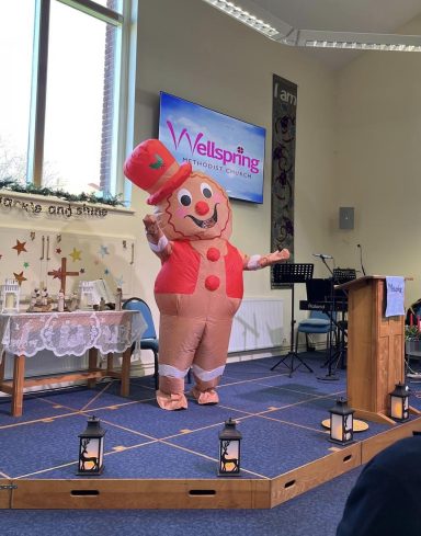 A gingerbread character in a festive outfit stands on stage, smiling and gesturing.